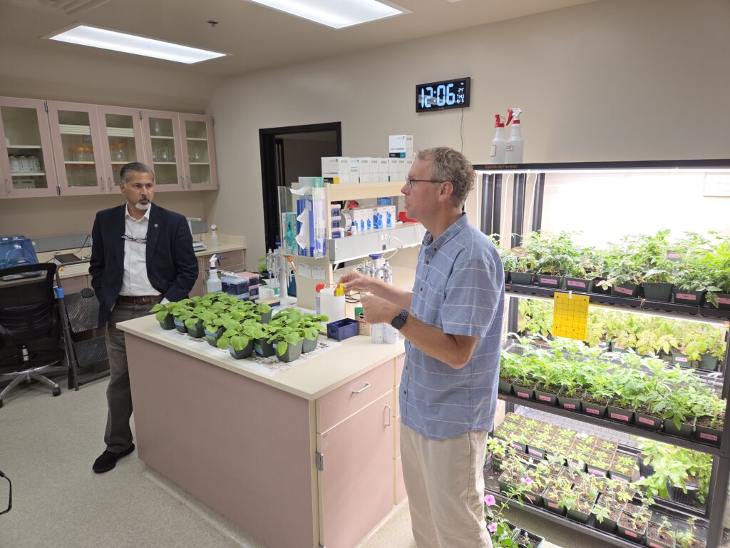 Raj Khosla and Rodney Cooper stand inside a room with plants on a countertop and many more plants on lighted shelves nearby. Cooper gestures while speaking; Raj looks on.