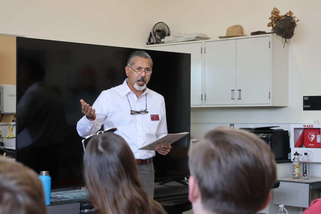 Raj Khosla gestures while speaking and holding a folder in one hand. Several people are seated, listening, with their backs to the camera.