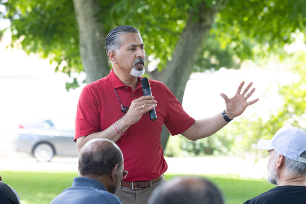 Raj Khosla gestures while speaking outside to a seated group with their backs to the camera.