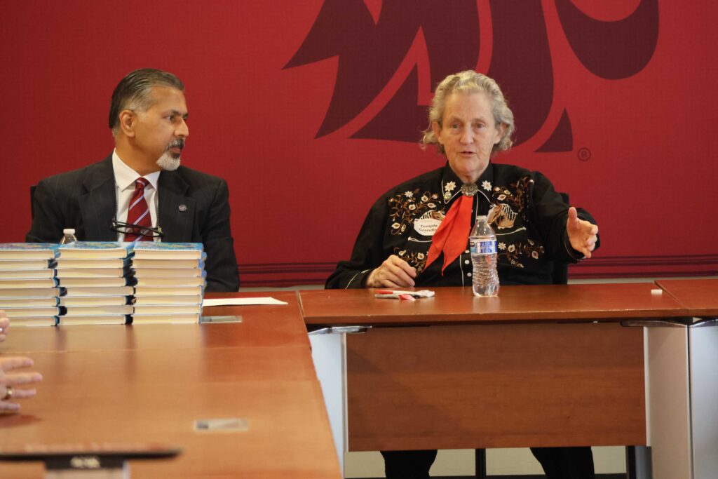 Raj Khosla looks on as Temple Grandin speaks. Both are seated at a table.