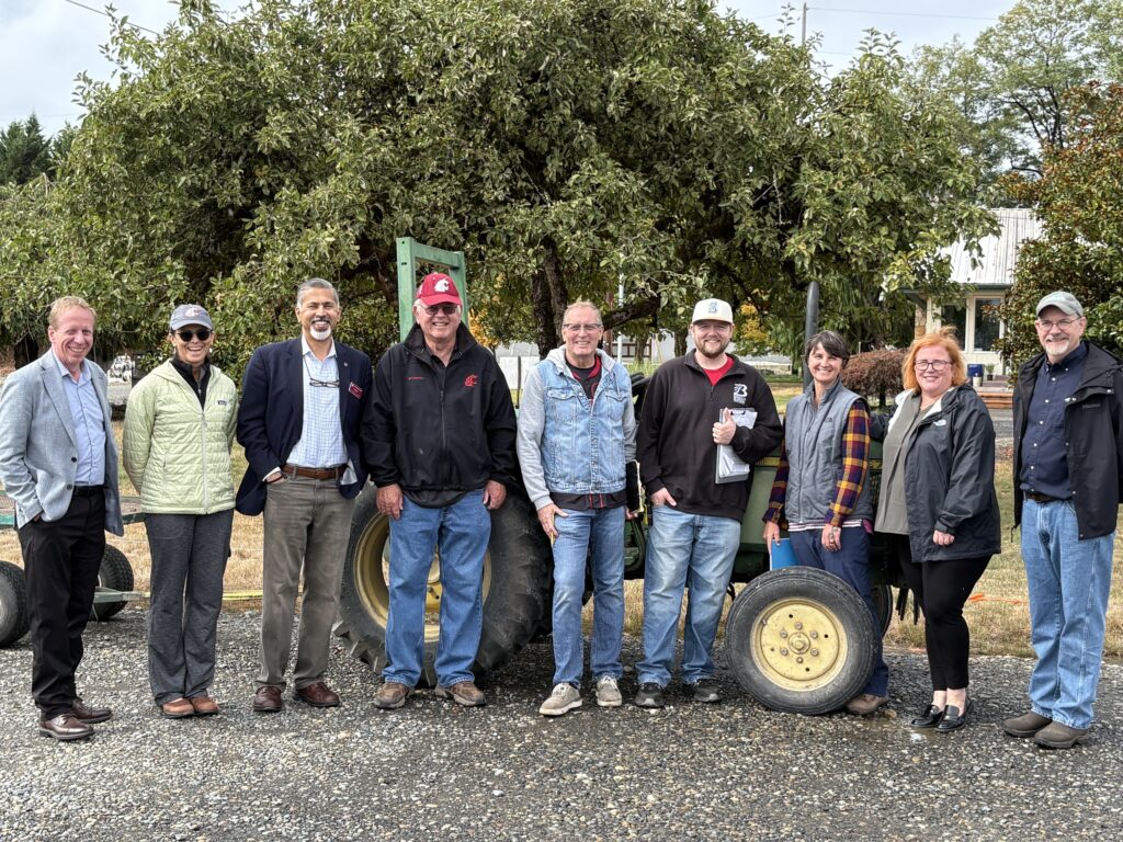A group photo of people standing near mature fruit trees with a tractor behind them. From left are a man in a gray sports coat; a woman in a light colored jacket and sunglasses; a man in a dark sports coat; a man in a black jacket and WSU cap; a man in a jean jacket; a man in a black jacket and Marines cap; a woman in a gray vest and flannel shirt; a woman in a black jacket; and a man in a dark blue shirt and black jacket.