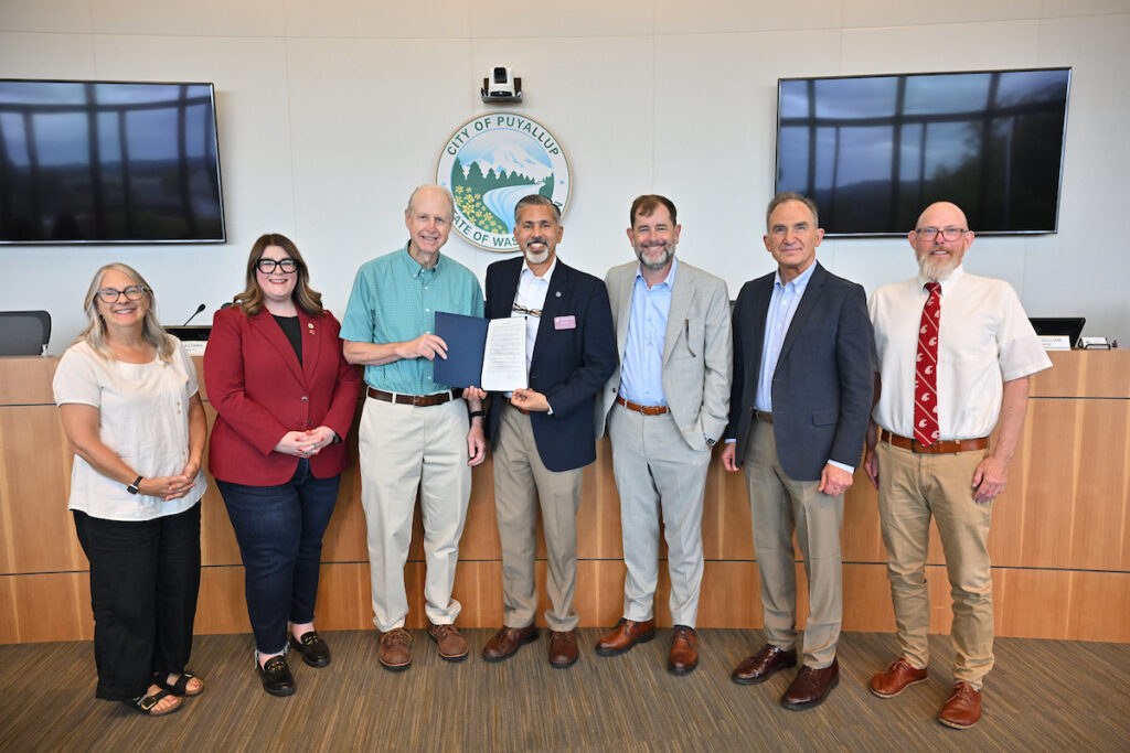 A group photo at a civic proclamation ceremony. From left are a woman in a white blouse; a woman in a crimson sports coat; a man in a teal button-down shirt, holding a document folder with a man in a dark sports coat; a man in a light gray sports coat; a man in a sports coat and light blue shirt; and a man in a white shirt and crimson tie. A wall seal behind them reads City of Puyallup.