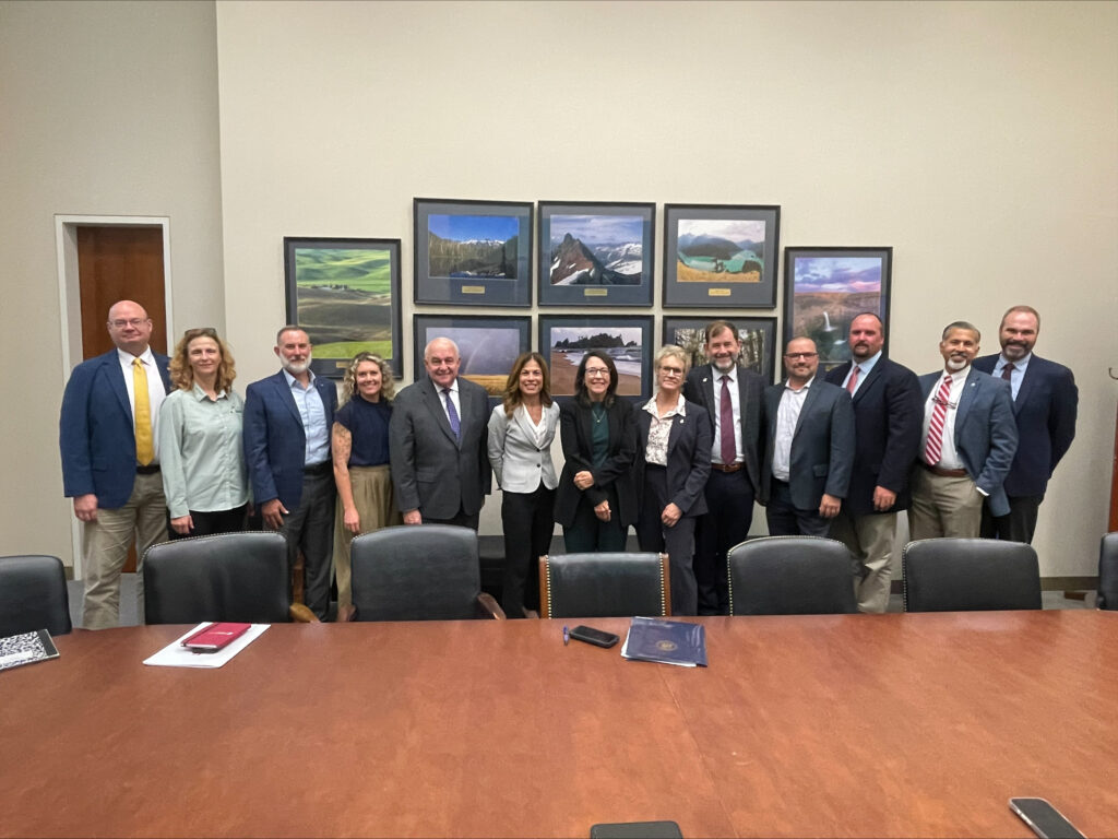 13 people stand side by side for a group photo in a conference room behind a large wooden meeting table. The room contains black leather chairs and framed landscape photographs arranged on the wall behind the group. Most individuals are wearing business or business-casual attire, including suits, blazers, collared shirts, and ties.