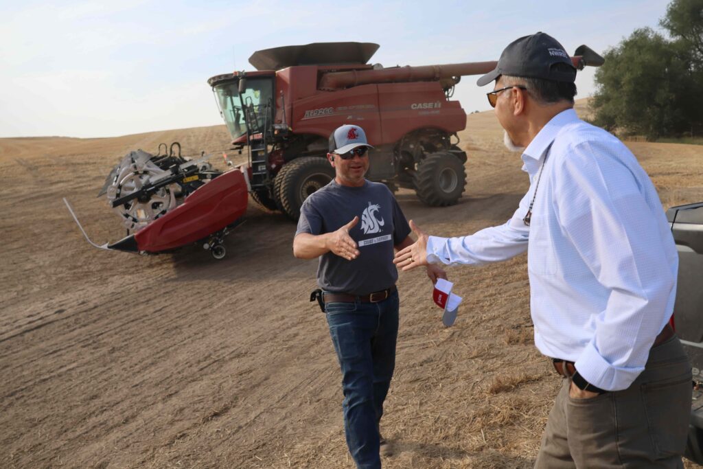 Raj Khosla and Kurt Druffel reach out to shake hands while standing next to a dirt field. In the background is a large piece of farm equipment.