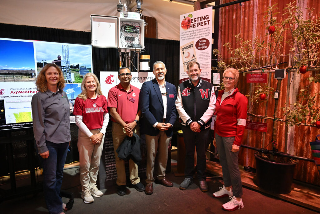 A group photo of visitors to a science exhibit. Standing are a woman in a gray shirt, a woman in a Washington State University shirt, a man in a crimson sports shirt, a man in a dark sports coat, a man in a WSU letterman's style jacket, and a woman in a crimson jacket. Exhibits behind them read AgWeather and Besting the Pest