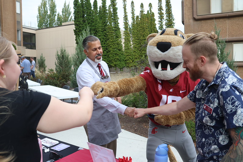 A female student facing away from the camera, a man in a white shirt, crimson tie, and apron, mascot Butch T. Cougar, and a man in a Hawaiian shirt and nametag exchange fist bumps near outdoor tables.