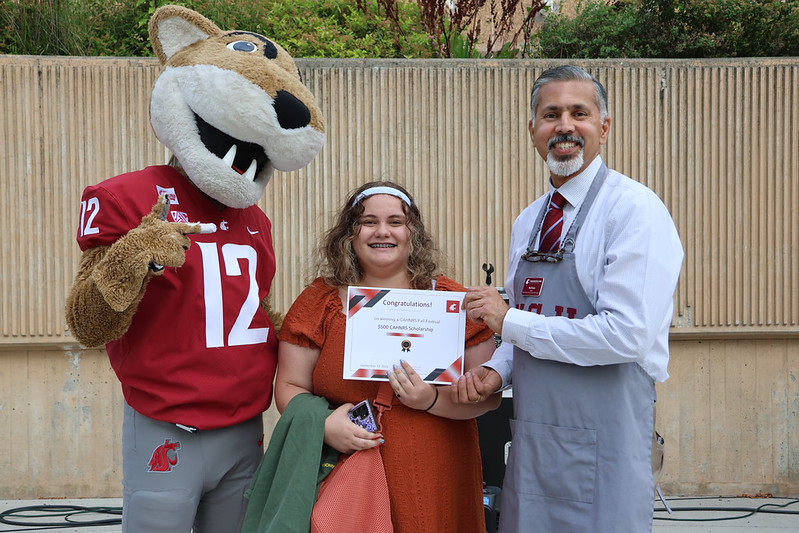 Butch T. Cougar, the WSU mascot, in a 12 jersey, gives a thumbs up as a woman in an orange dress and white headband holds up a scholarship certificate that reads Congratulations. Standing to her left is a smiling man in a white button-down shirt, crimson tie, and gray apron.