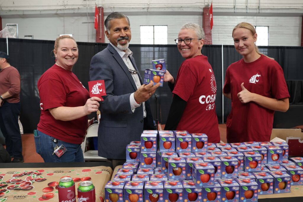 Four people stand behind two tables with many boxes of Cosmic Crisp® apples and Cosmic Crisp® stickers on them. One person holds a koozie with a white WSU cougar head logo on it. Two of the people hold boxed Cosmic Crisp® apples. The fourth person gives a thumbs up sign.
