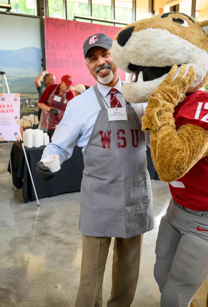 Raj Khosla holds a cup of ice cream while standing next to Butch, the WSU mascot. Raj is smiling while wearing a WSU apron and hat.