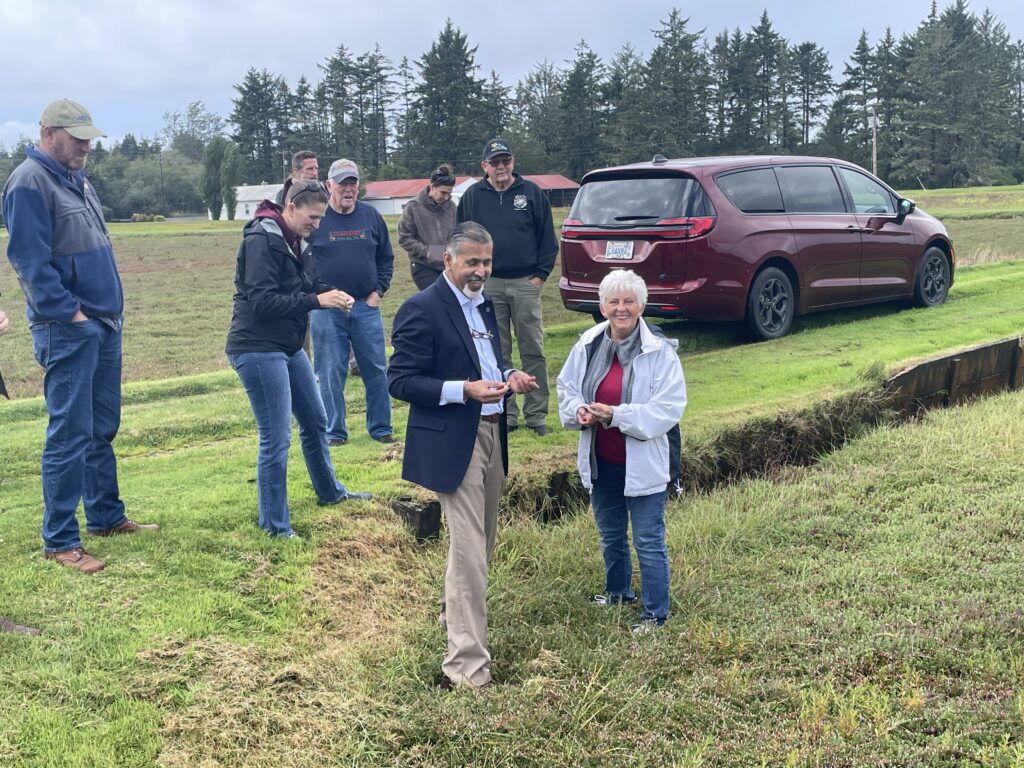 A man in a blue sport coat and light blue shirt stands with a woman in a light-colored jacket, holding small berries in their hands, as they stand in a sunken field. Behind them are several men and women, standing on a higher shelf of land near a maroon van. Farm buildings, a field, and trees are in the distance.