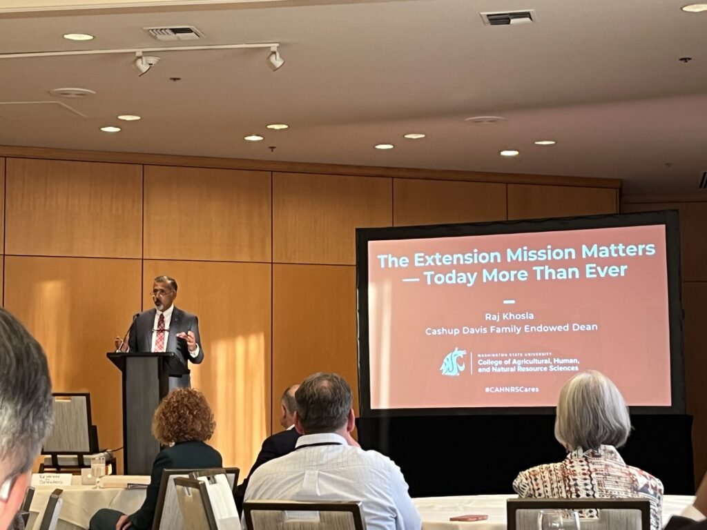 A speaker stands at a podium addressing an audience in a conference room with wood-paneled walls and recessed lighting. Attendees are seated at round tables facing a large presentation screen. The slide reads, “The Extension Mission Matters — Today More Than Ever,” with the presenter listed as Raj Khosla, Cashup Davis Family Endowed Dean, Washington State University College of Agricultural, Human, and Natural Resource Sciences. Near the text is the WSU cougar head logo. 