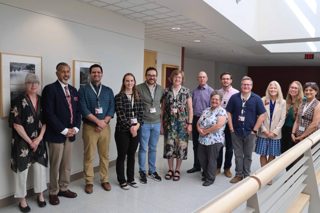 A group of 13 people poses for a photo in a bright university building hallway.