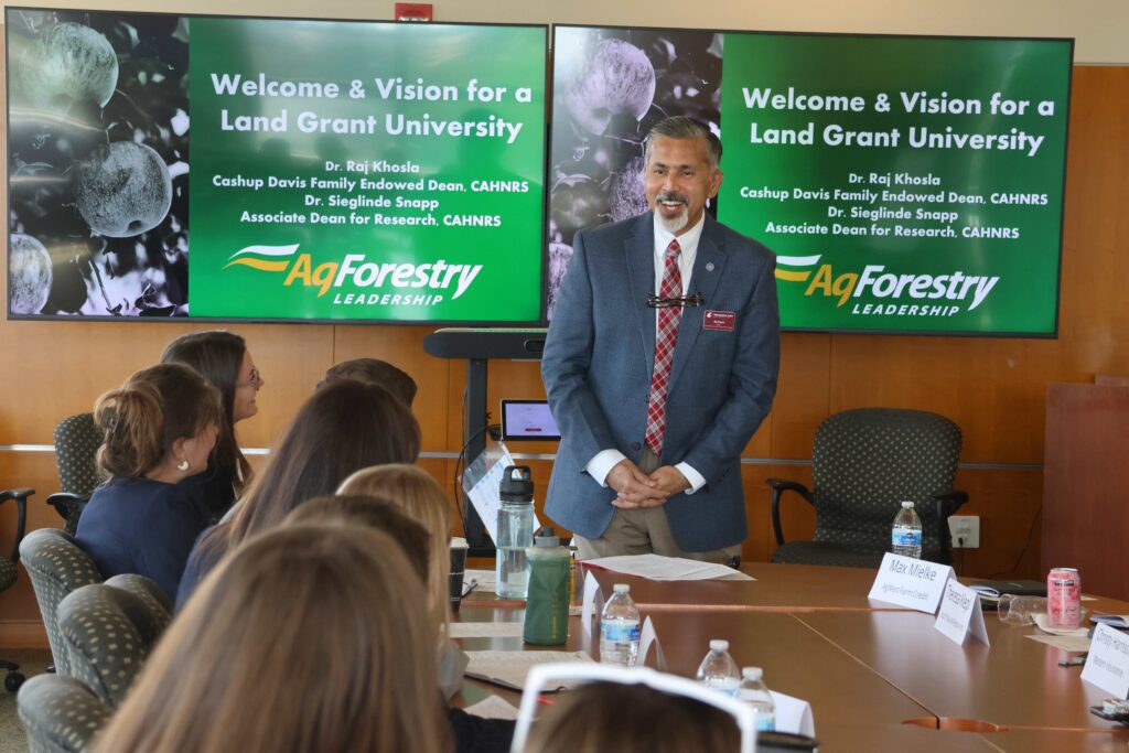 A man in gray suit and crimson plaid tie smiles as he speaks with a group of people seated facing him at a table. Behind him are screens reading "Welcome & Vision for a Land Grant University. Dr. Raj Khosla Cashup Davis Family Endowed Dean, CAHNRS Dr. Sieglinde Snapp Associate Dean for Research, CAHNRS AgForestry Leadership"