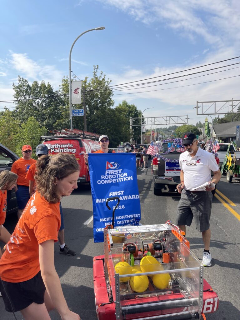 Participants in a community parade stand around a student-built robotics vehicle on a city street. One person holds a blue banner reading “FIRST Robotics Competition” and “FIRST Impact Award,” while several others wearing orange shirts gather nearby. The robotics machine, containing bright yellow balls, is displayed in the foreground. Decorated parade vehicles and floats line the street in the background, and a WSU banner hangs from a lamppost above the roadway.