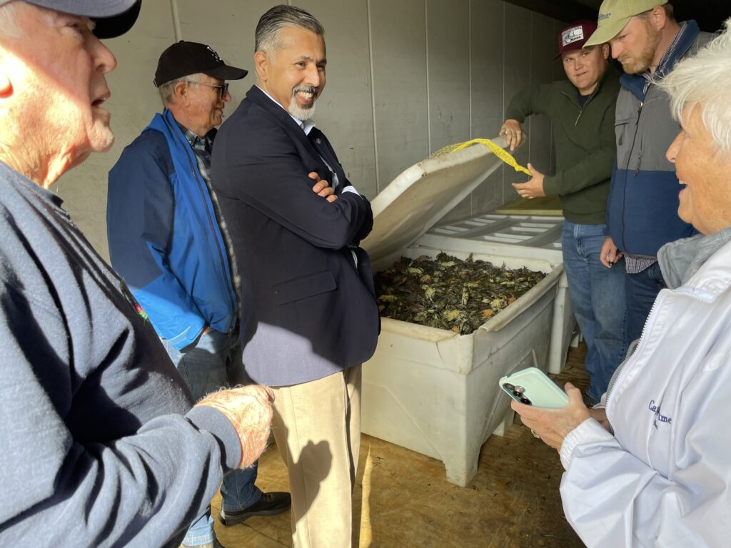 A man in a dark suit coat smiles as a group of men and women stand near a large bin of crabs. One man is holding open the bin's lid as others look on, talk, and smile. 