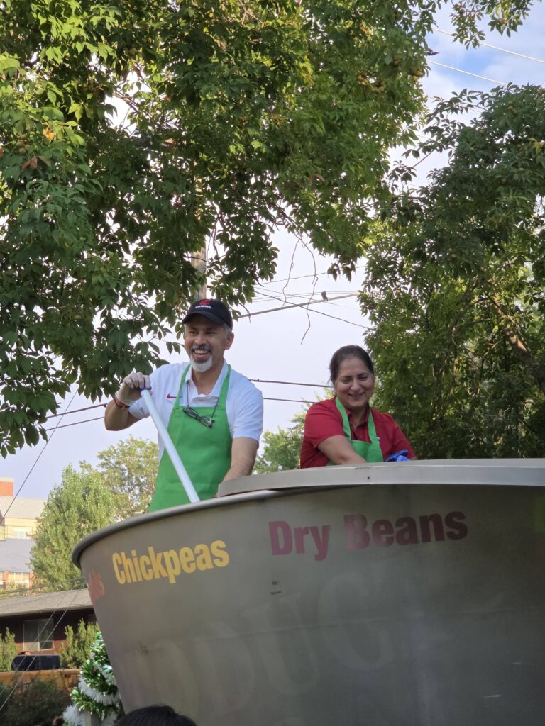 Raj Khosla and his wife stand next to a large cooking pot. Raj stirs the pot's contents. The side of the pot displays the words “Chickpeas” and “Dry Beans.” The pot is outdoors beneath leafy trees.
