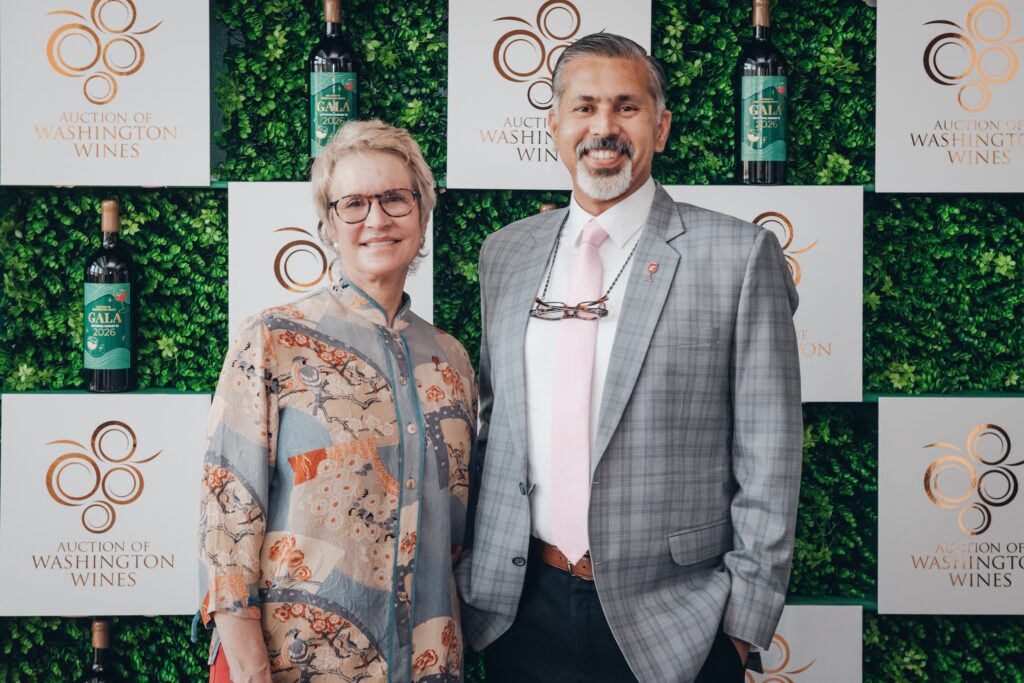 Raj Khosla and President Cantwell pose in front of an Auction of Washington Wines display featuring white panels with the Auction of Washington Wines logo, green foliage, and decorative wine bottles.