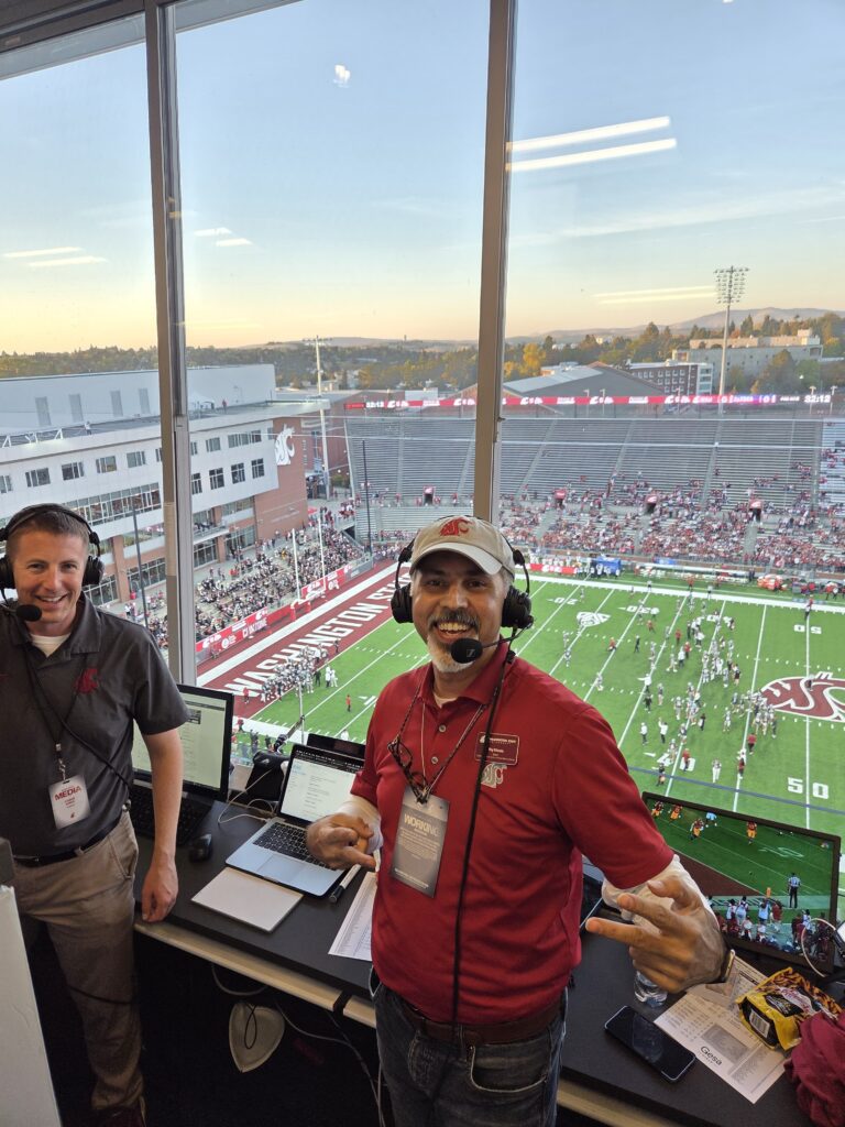 Raj Khosla gestures while standing next to another person inside a room with glass windows above the WSU football field. Both men are wearing headsets. On the desk behind them are several computers. 