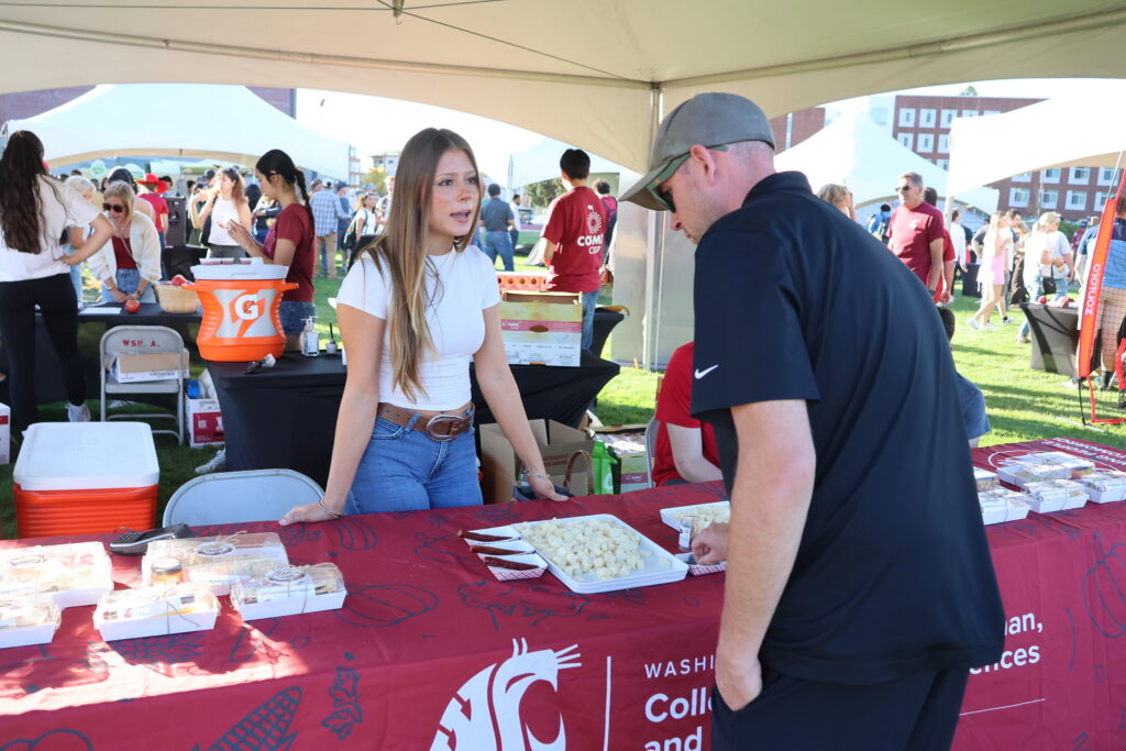 A person stands behind a table with a WSU CAHNRS-themed tablecloth on it. Another person stands in front of the table. On the table is a tray of cheese samples, beef stick samples and other food items. The table is under a tent. Several people can be seen in the background.