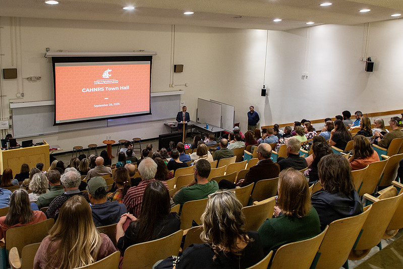 The backs of a large crowd seated in an auditorium. Dean Khosla stands at a podium at the front of the room. A projector screen at the front of the room reads "CAHNRS Town Hall" with the WSU cougar head logo on it.