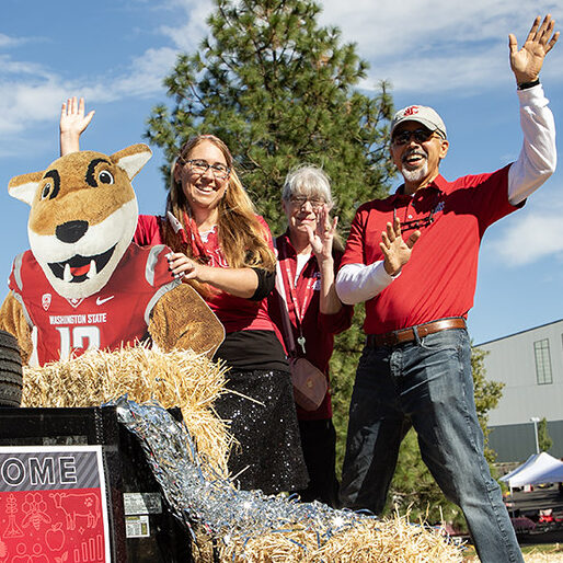 Three people wave from a parade float that has hay and a cutout cardboard replica of Butch the WSU cougar mascot.