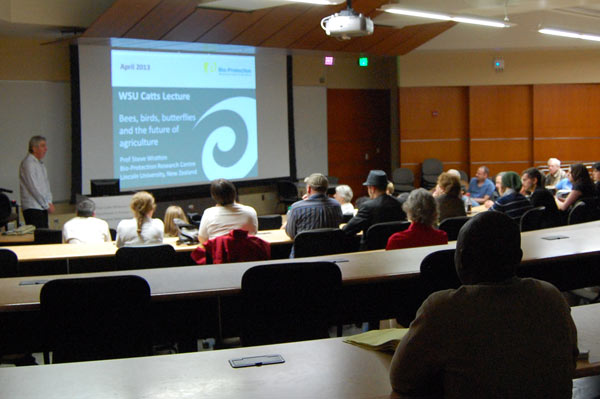 A man stands next to a projector screen that says, "WSU Catts Lecture. Bees, birds, butterflies, and the future of agriculture." People are seated and listening to the licture.