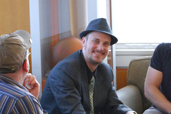 A man wearing a fedora smiles.