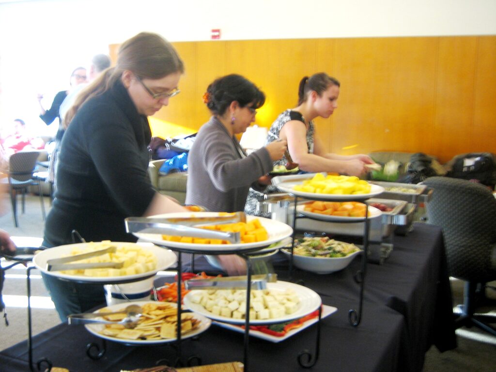 Various food items are laid out on plates on a table. People fill their own plates with food.