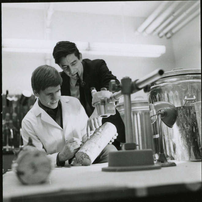 An entomology student and professor are working in a lab together. The professor stands smoking a pipe, pointing out something in a log sample.