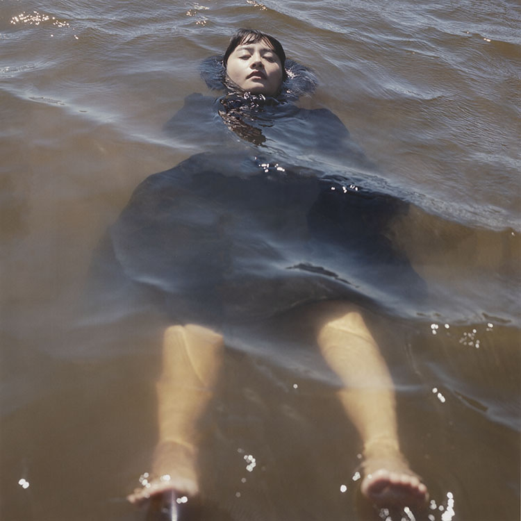 Photograph of woman in black dress floating on her back in brown choppy water. 