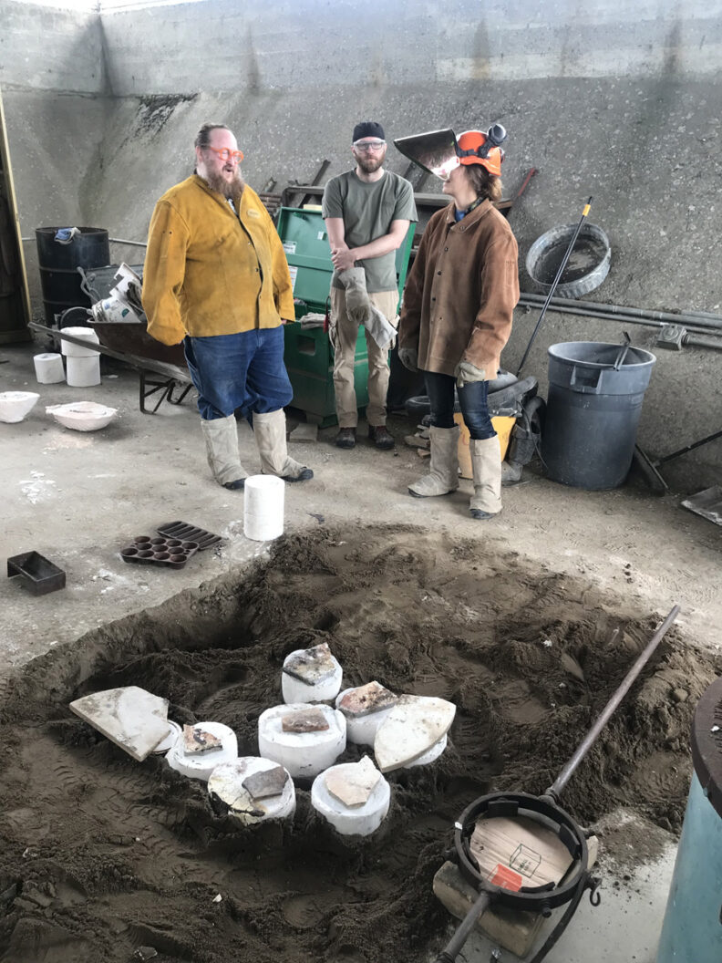 Artists standing by sandy pit with sculptures. 