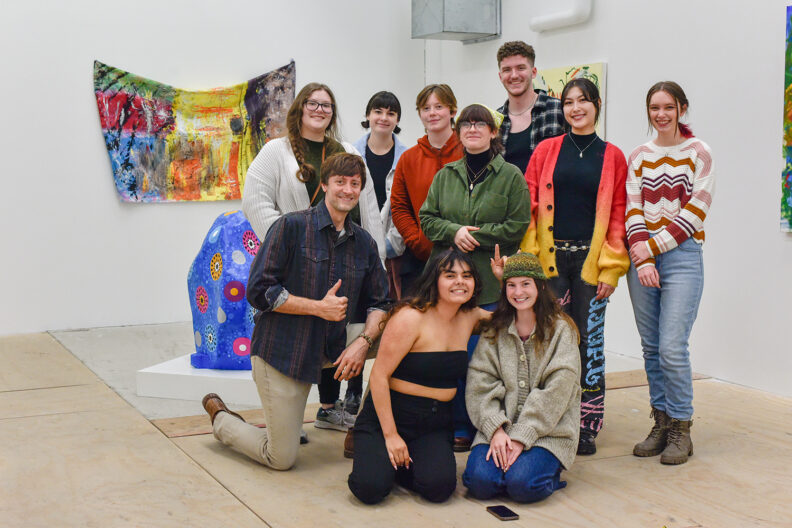 Group of art students posing in Locker Gallery with their painting professor and their work. 