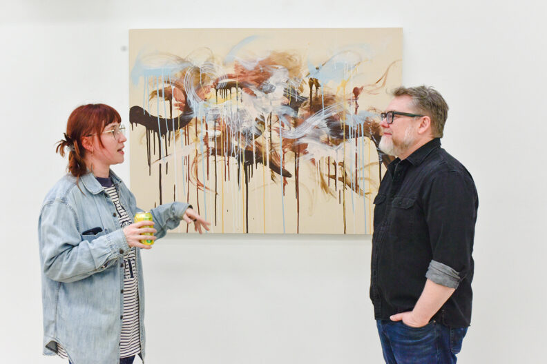 Artist in front of painting in gallery discussing work with instructor. 