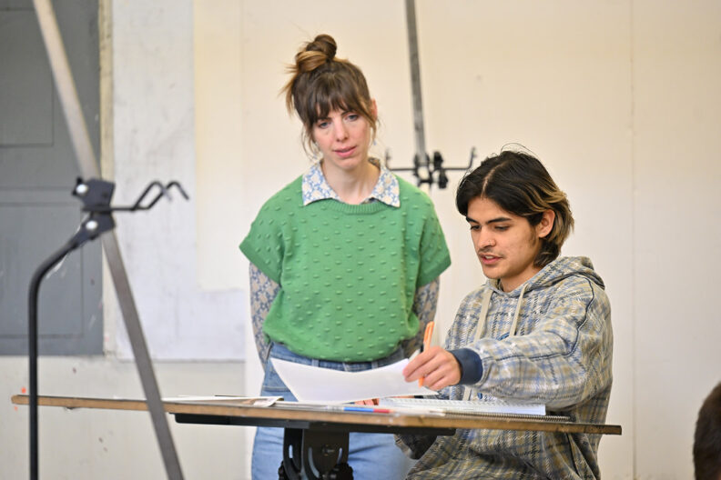 Art instructor speaking with student working at drawing desk.