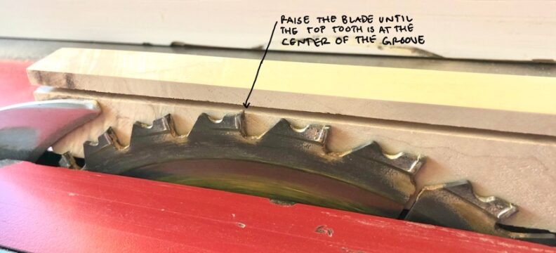 Photo of a raised table saw blade, with the material behind it. The groove that was cut in the previous step is visible above the blade. Instructions to raise the blade until the top tooth is at the center of the groove. Image links to larger version.