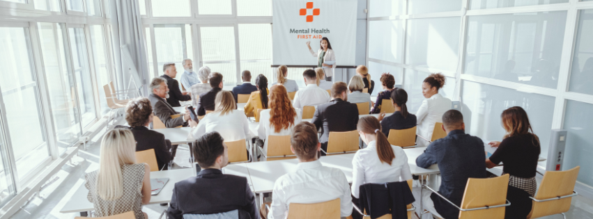 Group of people sitting in a board room at tables, listening to a presentation.
