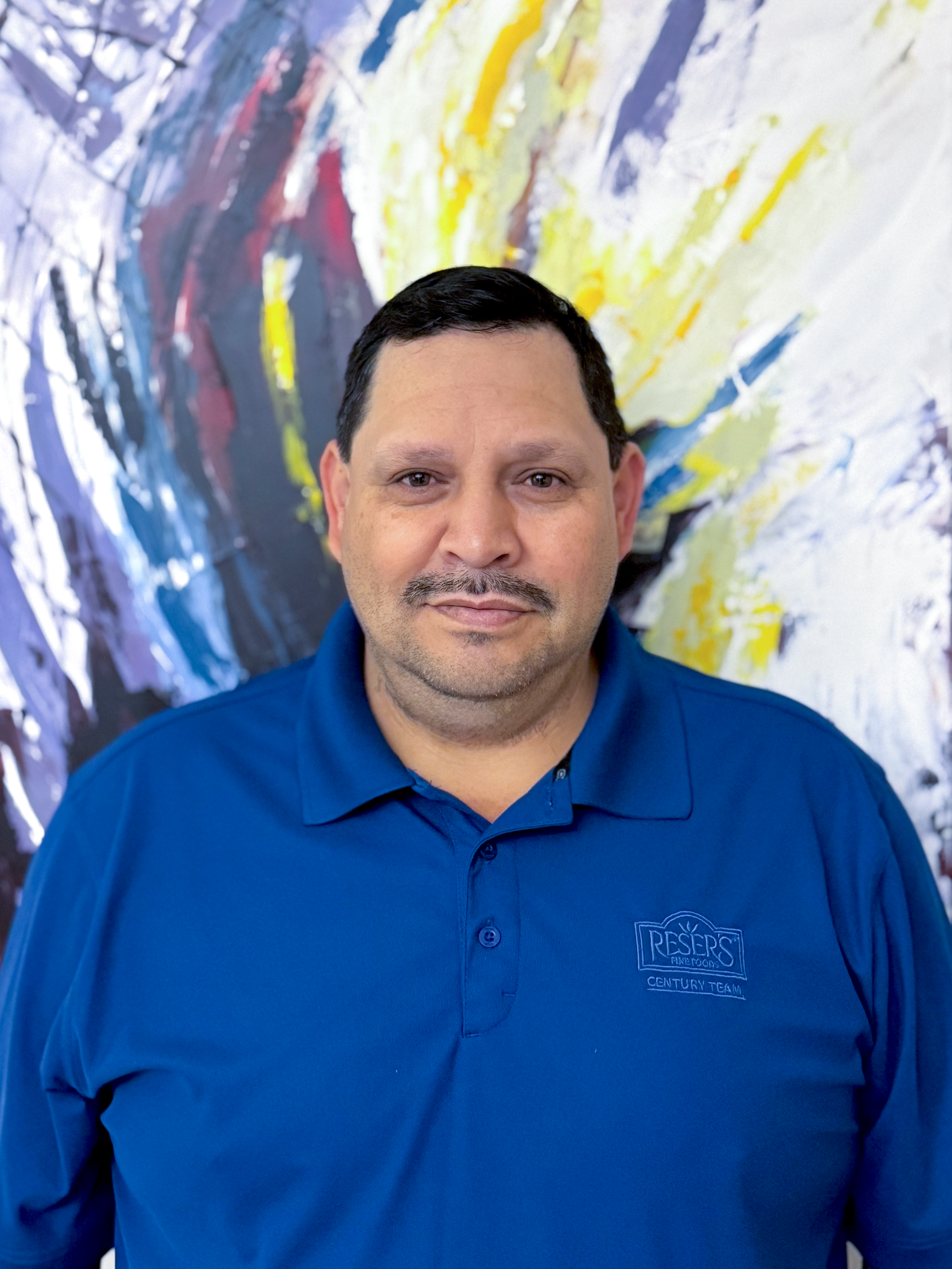 Jose Angel-Nolasco standing next to a colorful painting.  He works at Reser's fine foods