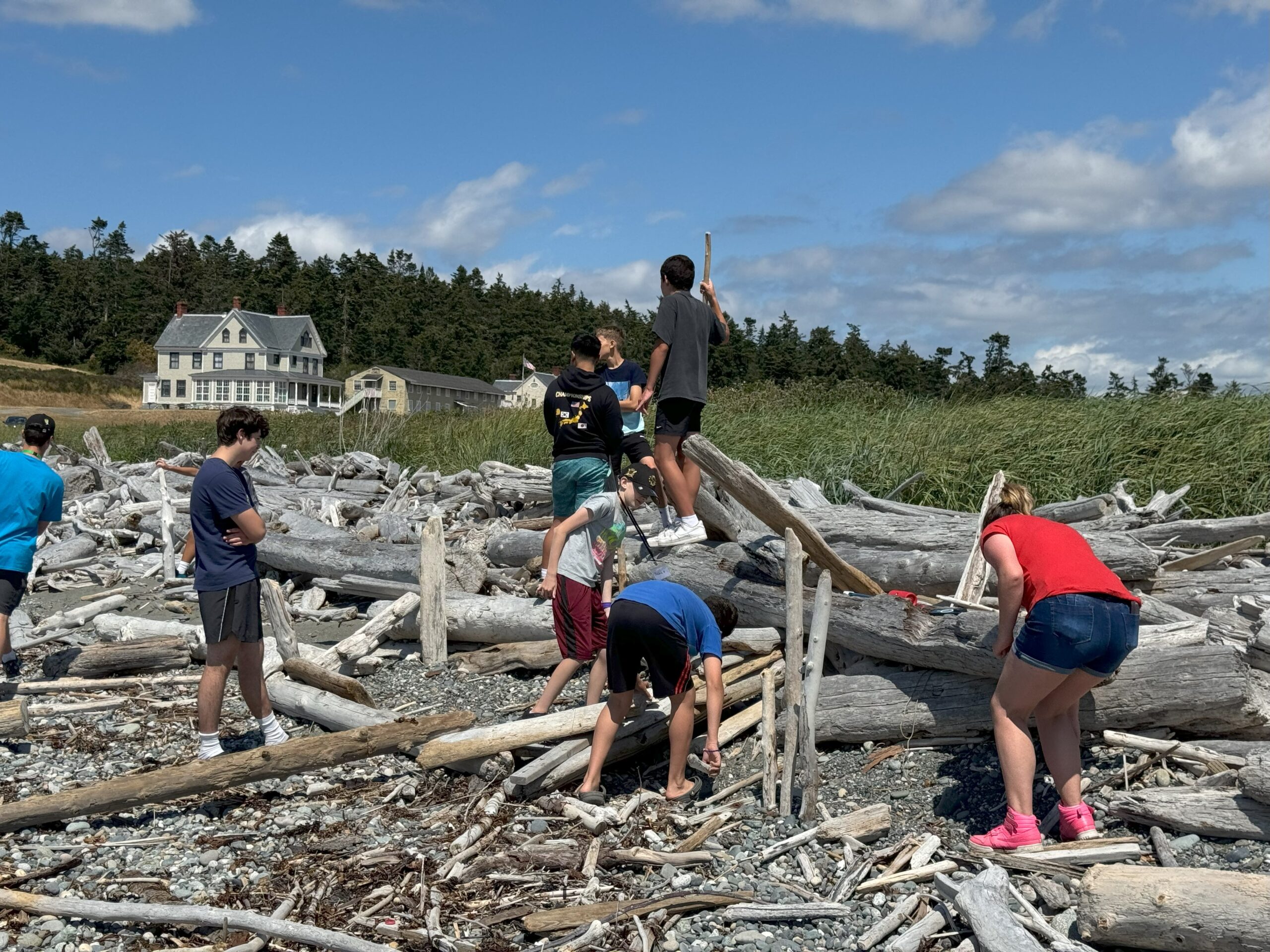 group of youth on the beach, looking at the driftwood, with Camp Casey in the background