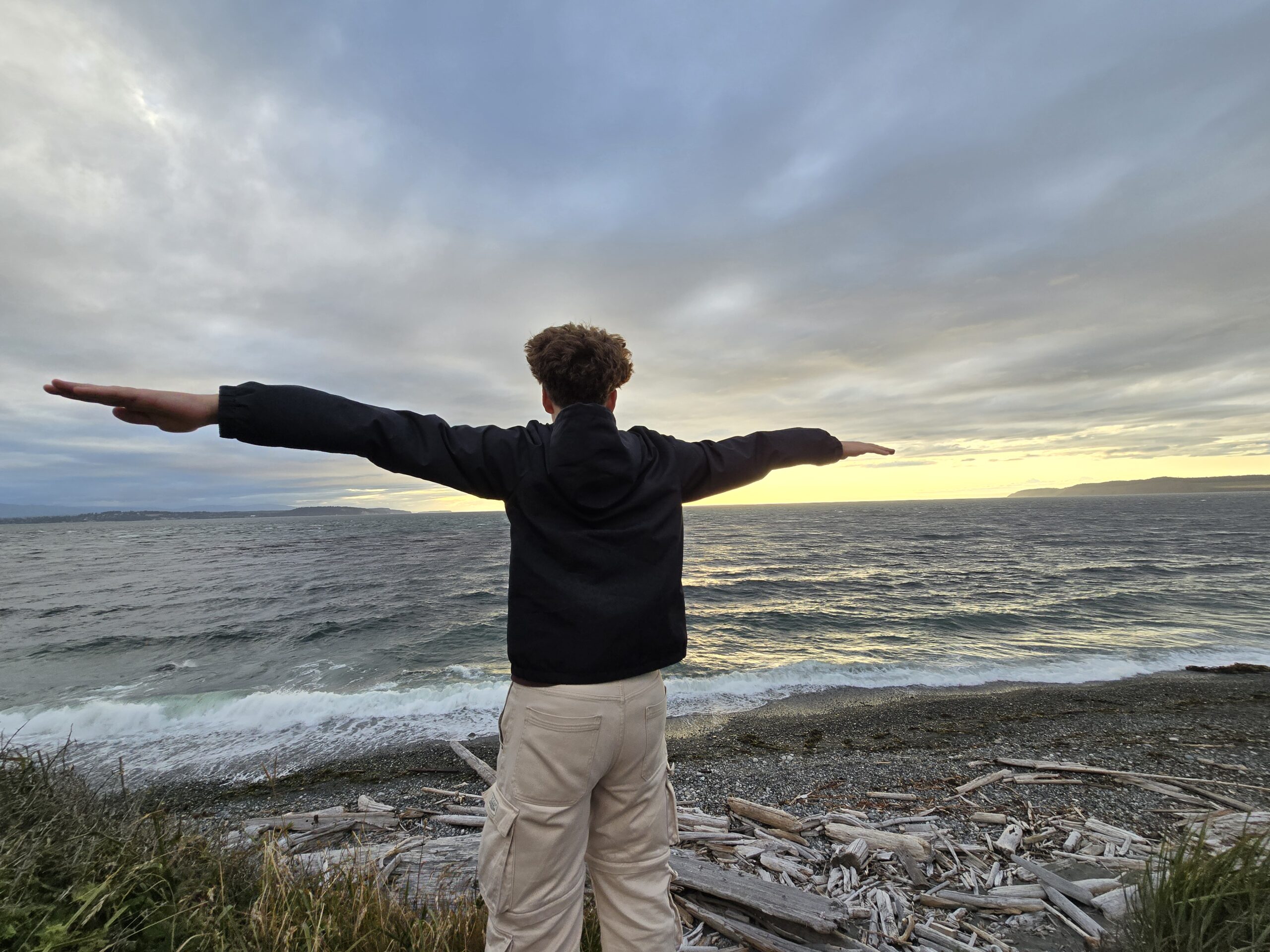 young man on the beach with arms outstretched facing away from the camera and towards a sunset.