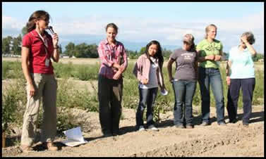 Six people standing beside a field; one woman is speaking with a microphone.