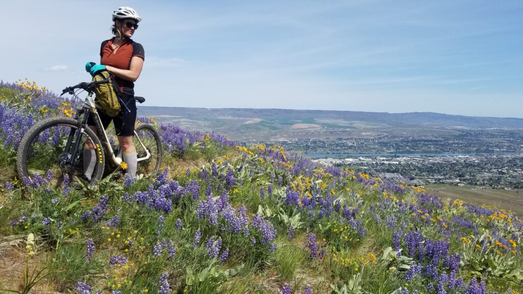 Woman on a mountain bike standing on a flower covered hill with wenatchee in the valley below.