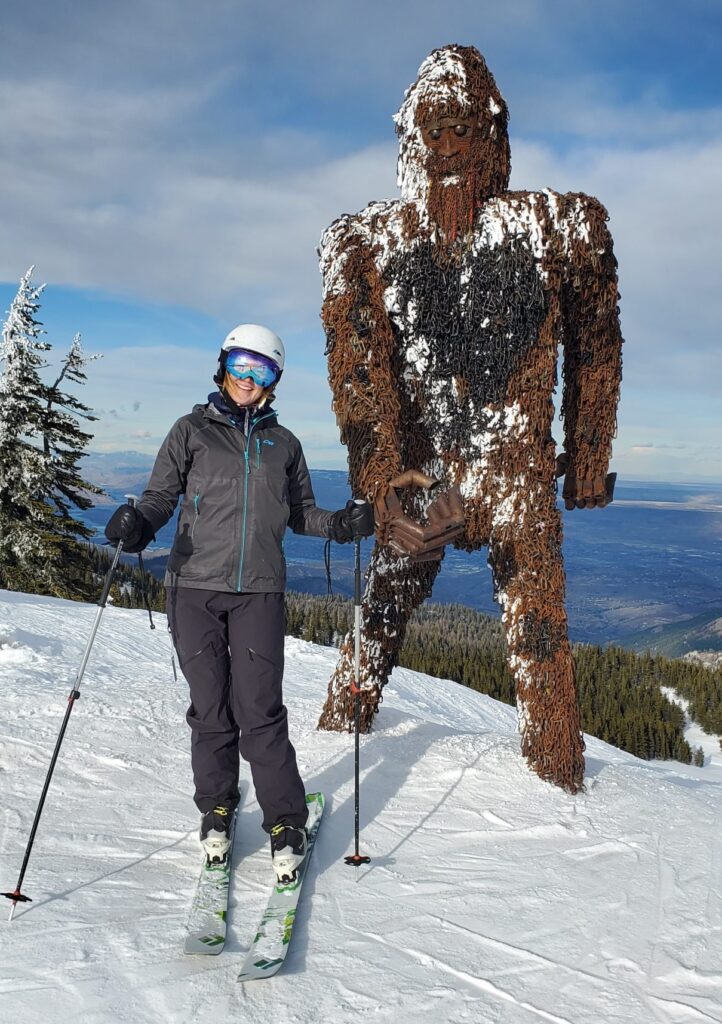 woman on skis standing in front of a very tall yeti sculpture at the top of mission ridge ski area.