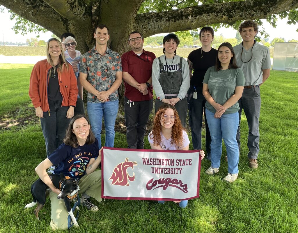 a group of lab members next to the trunk of a very large tree with outward branches above their heads. the two members in the front are kneeling and holding a WSU cougars banner between them. the one on the left is also holding a very cute black and white dog