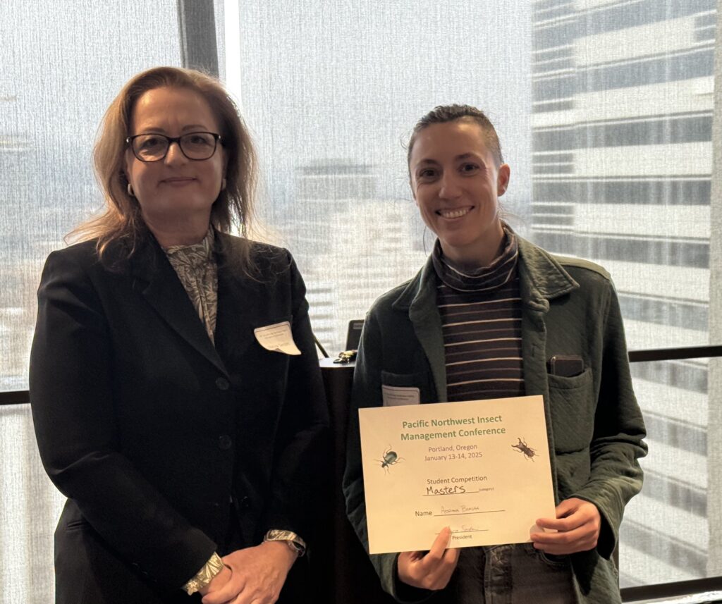 two people standing in front of a high-rise office window that shows Portland in the background. Adriana Barsan on the right is holding a certificate for winning the graduate student talk competition at the Pacific Northwest Insect Management Conference