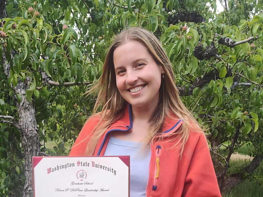 A smiling young woman with long light brown hair and and orange jacket worn over a light blue tee shirt is holding a certificate from Washington State University