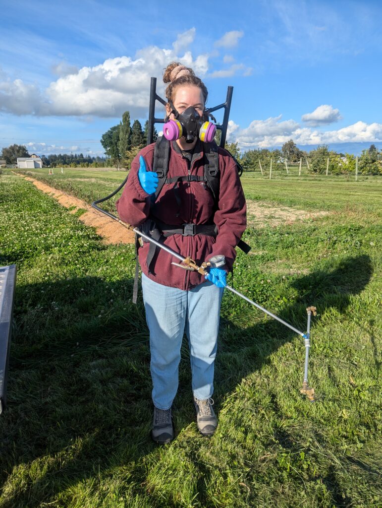 a person wearing a blue nitrile gloves, a respirator and a backpack sprayer holds the sprayer wand in one hand and is giving a thumbs up with the other hand.