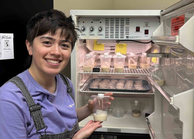 A smiling person with short dark hair wearing a purple tee shirt and overalls is holding a container of foamy liquid in front of an open lab fridge containing more containers of foamy liquid.
