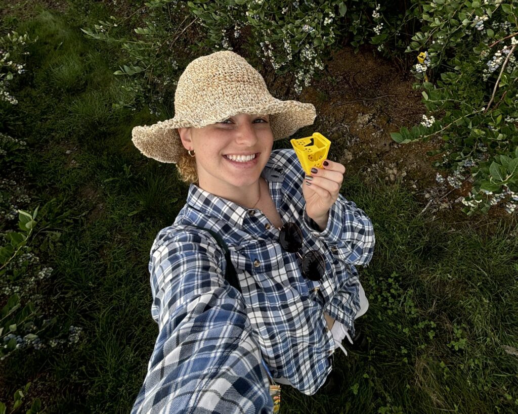 Claire, looking fabulous in a straw hat and plaid shirt is against a green leafy background holding a yellow decoy dispenser