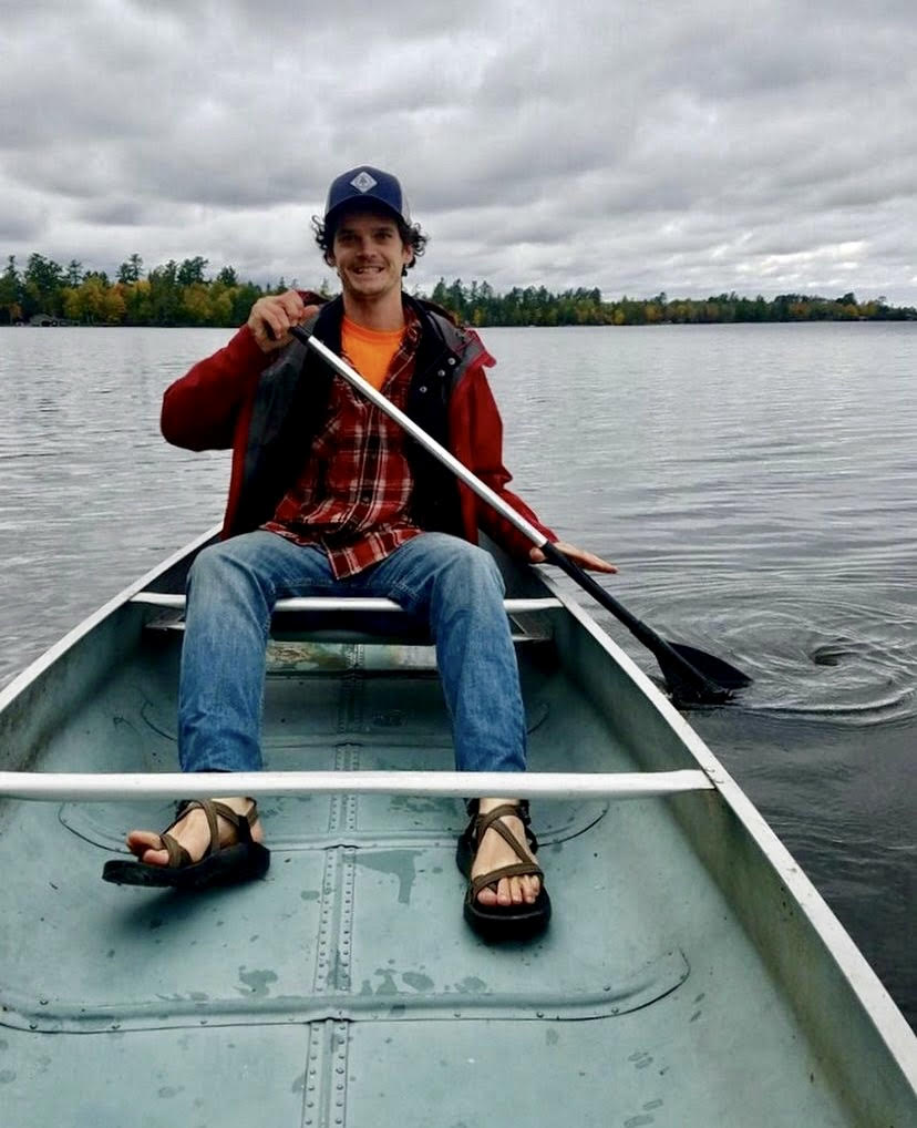 man in a red sweatshirt and baseball cap sitting in an aluminum canoe paddling on a lake