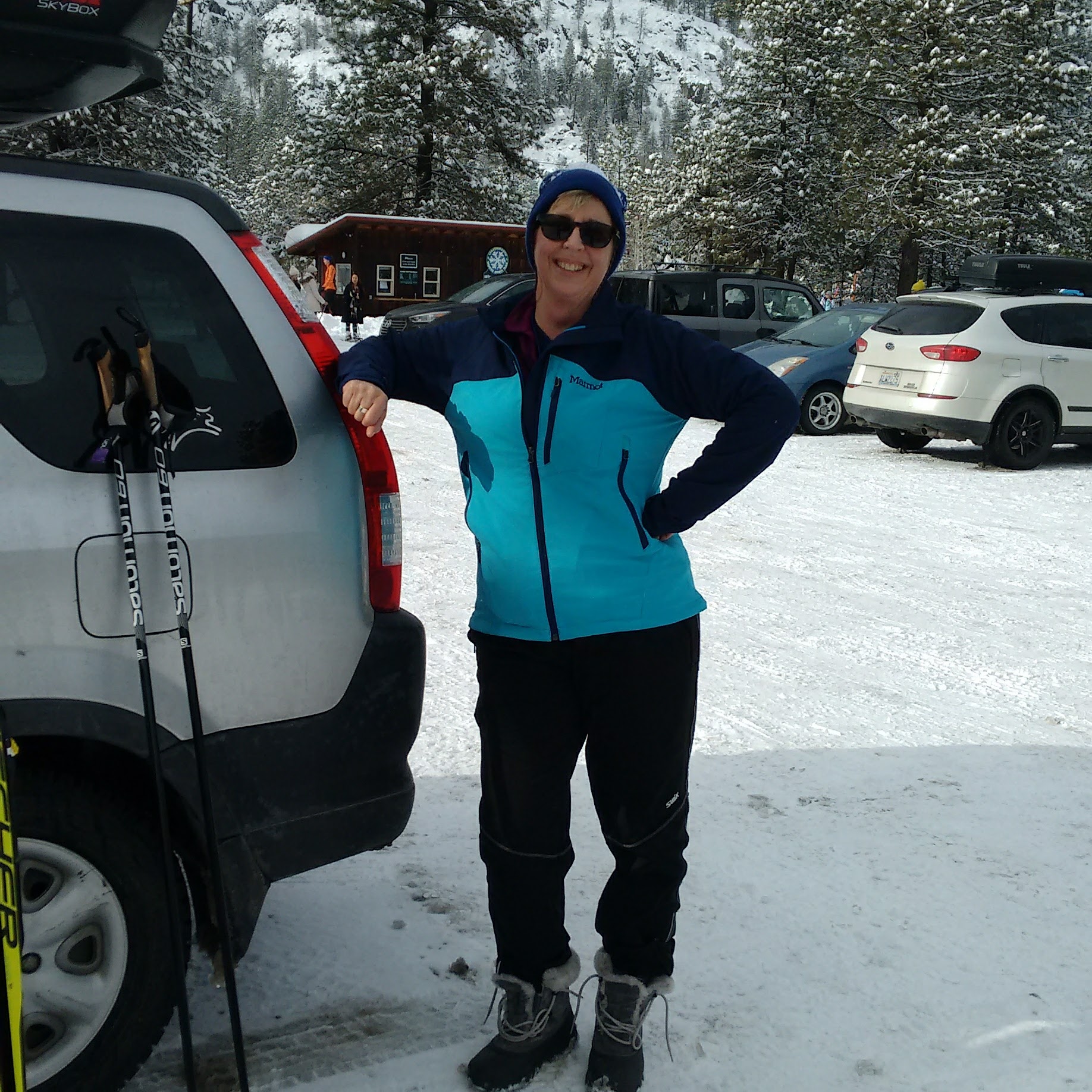 a smiling woman wearing a blue and teal jacket and matching hat and sunglasses is leaning on a silver car in a snowy parking lot